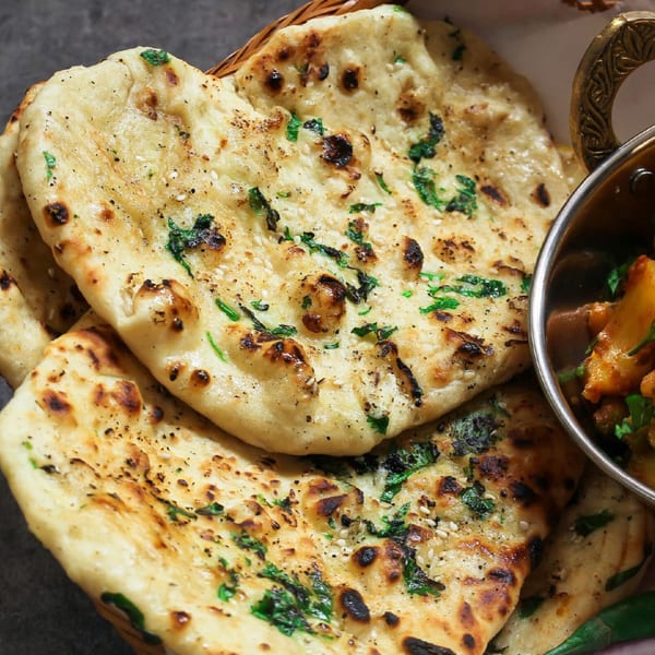 Close-up of golden-brown naan bread with cilantro and sesame seeds, next to a bowl of Indian curry.