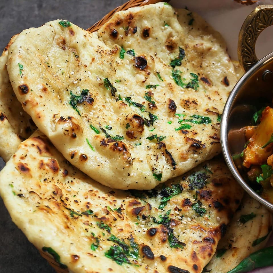 Close-up of golden-brown naan bread with cilantro and sesame seeds, next to a bowl of Indian curry.